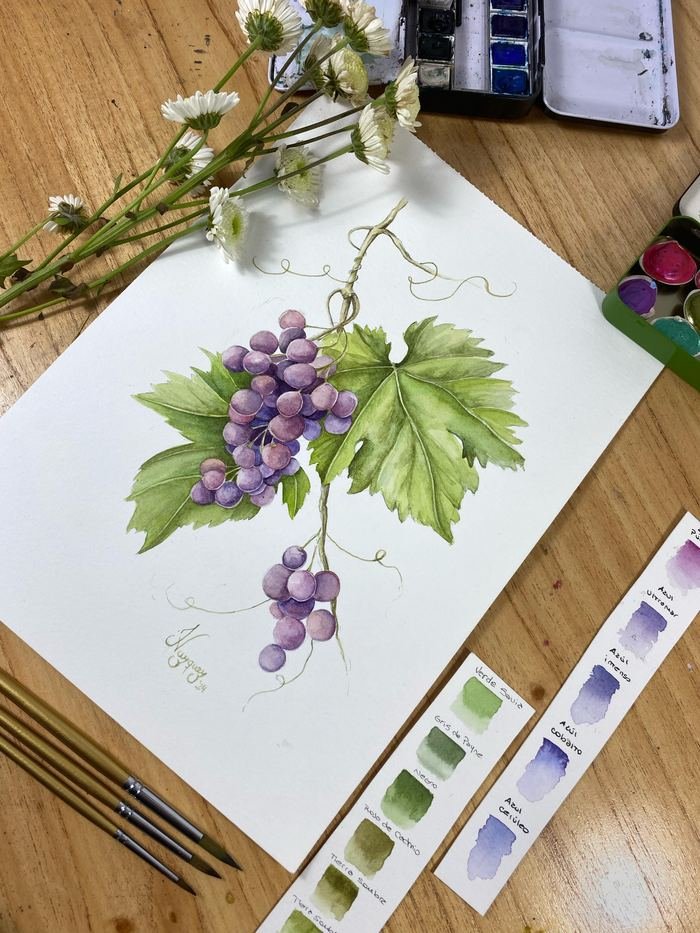 Acuarela de un racimo de uvas moradas con hojas de parra, sobre la mesa de trabajo con los pinceles y las muestras de color