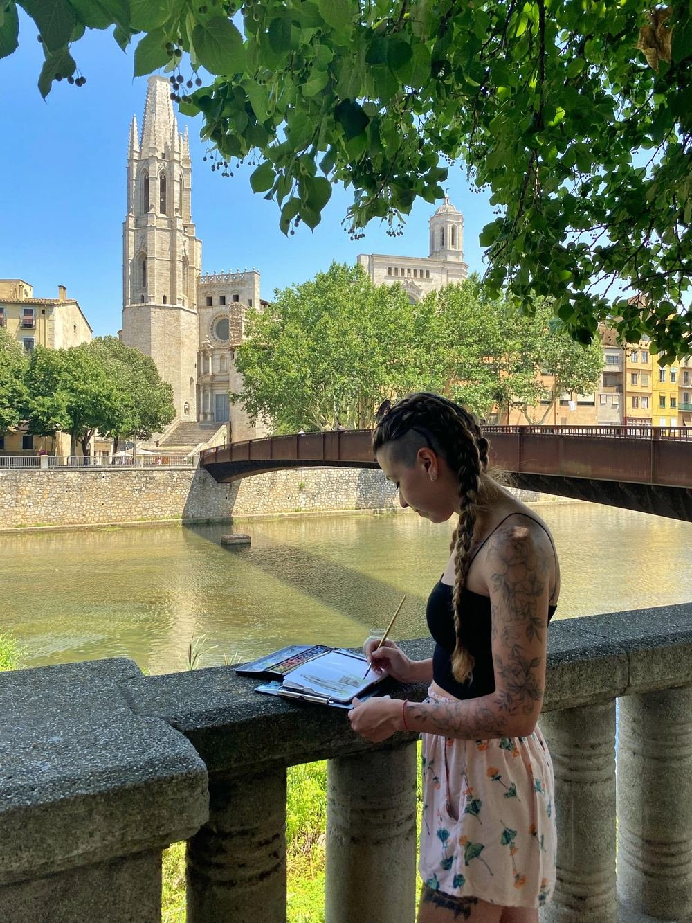 Julieta dibujando en su cuaderno de viaje, apoyada en una baranda de piedra frente a un río, con una catedral de fondo