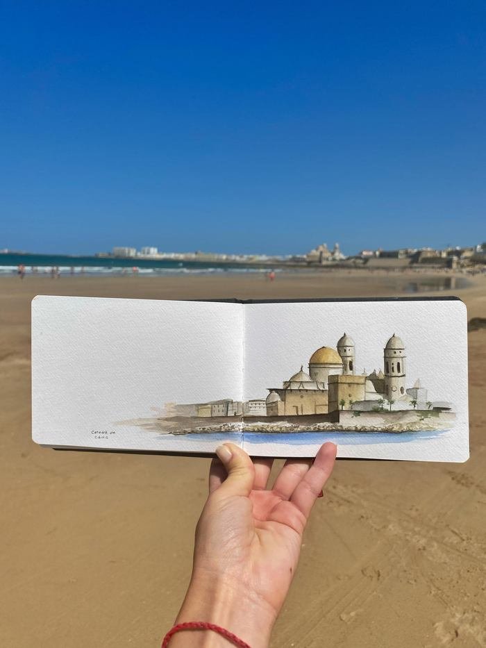 Sketchbook con la catedral de Cádiz en acuarela, sostenido en la arena frente al mar